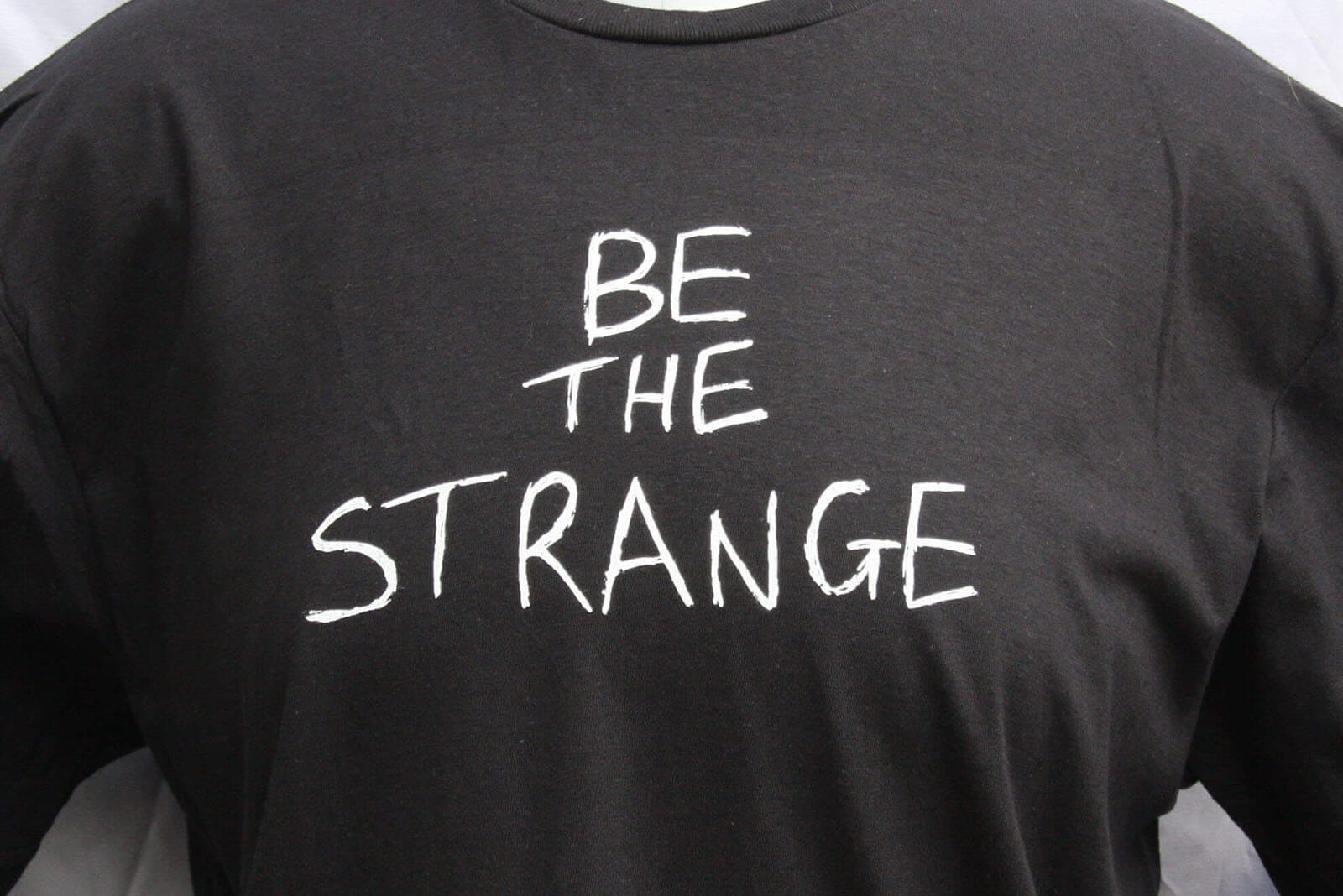 Black t-shirt with 'BE THE STRANGE' text in white on a plain background ElRat Designs