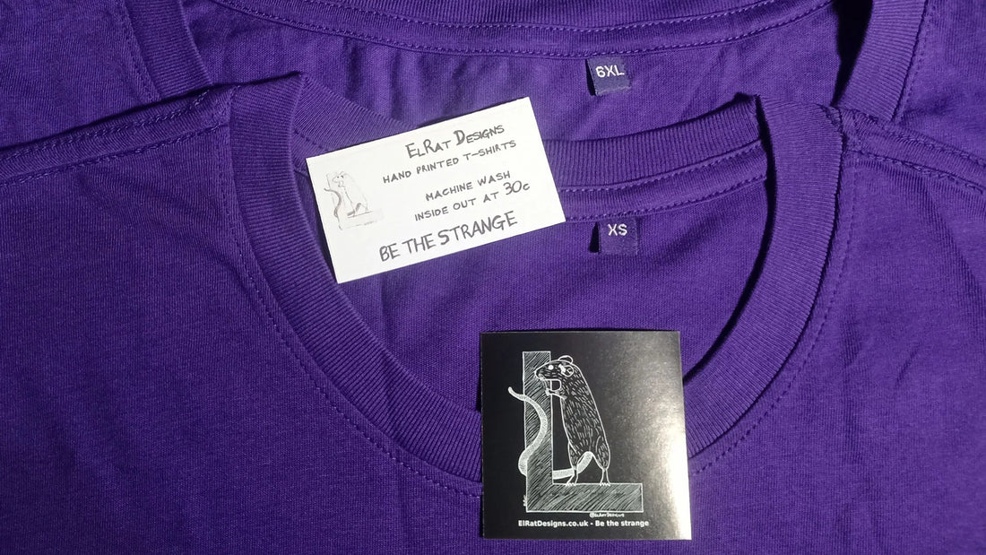 Purple ElRat Designs t-shirt showing XS and 6XL size tags with wash card reading "Be the Strange" - inclusive sizing from extra small to 6XL