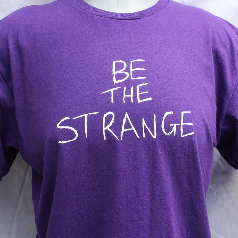 Purple t-shirt with 'BE THE STRANGE' text on a plain background ElRat Designs