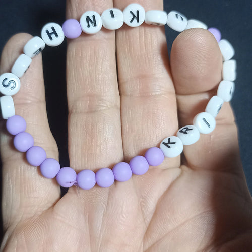 Bracelet with letter beads spelling out 'KRISTIN FN HERSH' on a hand against a dark background, Pale Purple