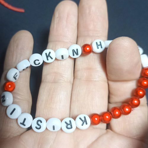 Bracelet with letter beads spelling out 'KRISTIN FN HERSH' on a hand against a dark background, Red