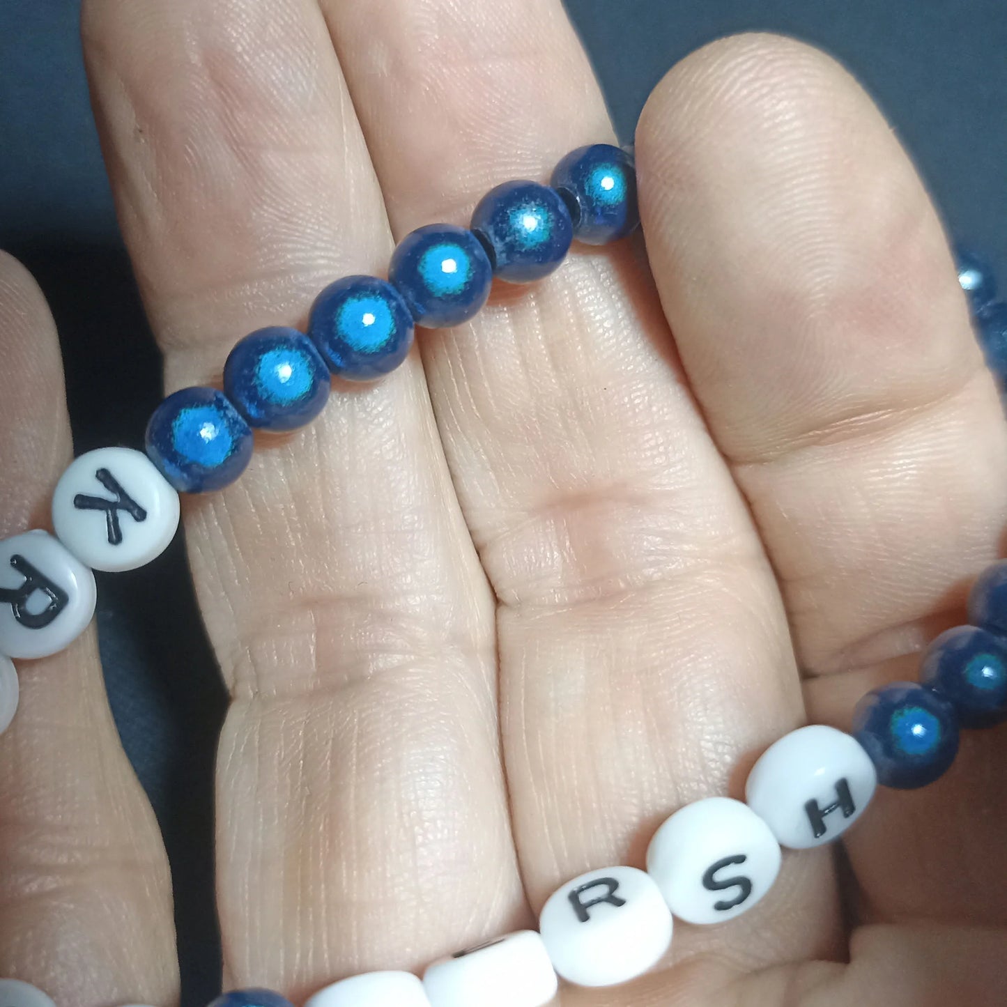 Blue beaded Kristin Hersh bracelet with white letter beads on a hand against a dark background, Clear Dark Blue