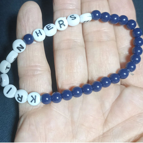 Blue beaded Kristin Hersh bracelet with white letter beads on a hand against a dark background, Dark Blue