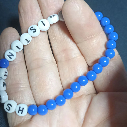 Blue beaded Kristin Hersh bracelet with white letter beads on a hand against a dark background, Opaque Mid Blue