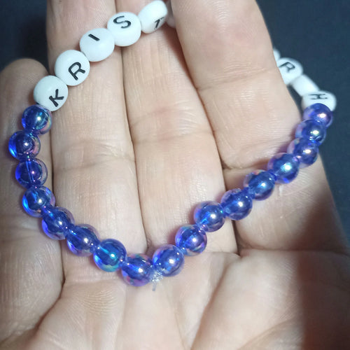 Blue beaded Kristin Hersh bracelet with white letter beads on a hand against a dark background, rainbow mid Blue