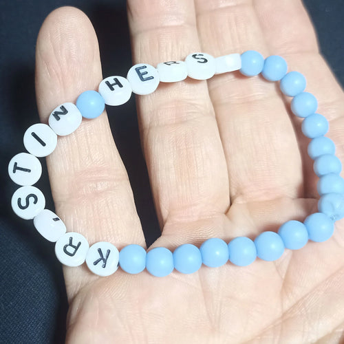 Blue beaded Kristin Hersh bracelet with white letter beads on a hand against a dark background, Opaque Pale Blue